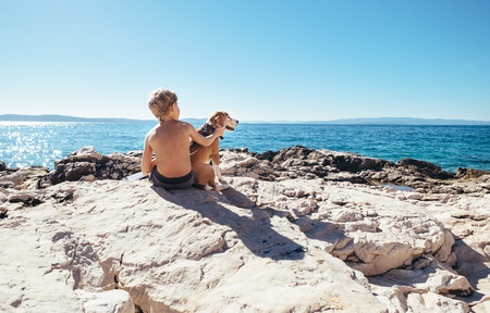 Boy with beagle dog sitting together on rocky sea coast at sunny afternoonの写真素材