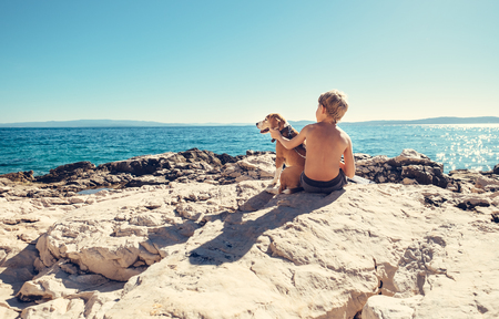Boy with beagle dog sitting together on rocky sea coast at sunny afternoonの写真素材