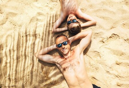 Father and son lie togetther on the sand under the palm tree and take a sun buthの写真素材