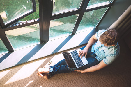 Man works with laptop sitting on the floor near the open windowの写真素材