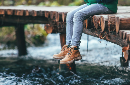 Boy sits on the wooden bridge over the river. Close up legs in boots image