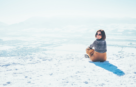 Happy smiling woman sits on the snowy mountain hill over the city. Sunny winter beautiful weather dayの写真素材