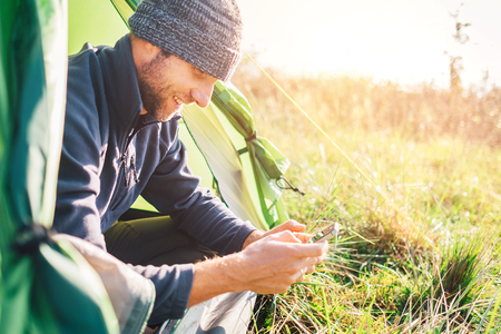Man traveler portrait using his mobile phone and sitting in camping tent in wild natureの写真素材