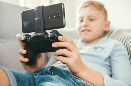 Preteen boy use gamepad with smartphone plays games lying on the cozy sofa in the home living room.の写真素材
