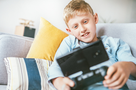 Preteen boy plays with smartphone connected with gamepad lying on the cozy sofa in the home living roomの写真素材