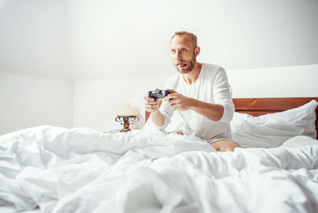 Big little boy: adult man waked up and plays PC games sitting in bedの写真素材