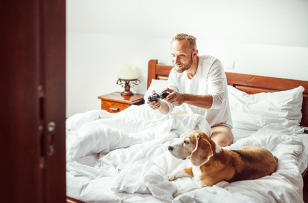 Man plays PC games sitting in bed at morning time. His beagle dog watching the game with very interest.の写真素材