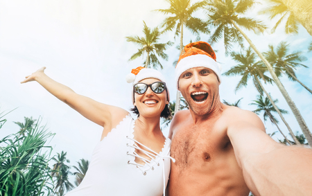 Cheerful young people couple dressed red Santa hats making selfie with tropic palm treesの写真素材