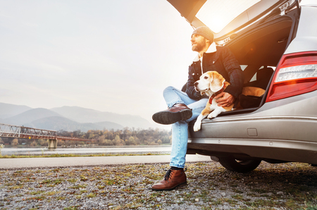 Breaded man in warm clothes siting with beagle in car trunk. Traveling with petの写真素材