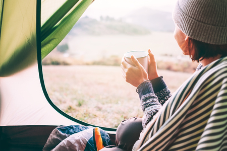 Happy female with cup of hot tea sits in camping tent, looks out and enjoys a new day beginningの写真素材