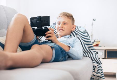 Preteen boy use gamepad with smartphone plays games lying on the cozy sofa in the home living room.の写真素材
