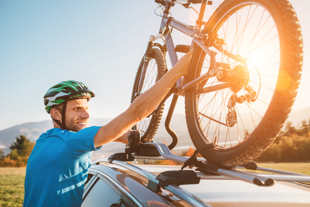 Young Man veared in cycling clothes and protective helmet instals his mountain bike on the car roofの写真素材