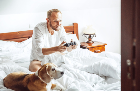 Adult man waked up and plays PC games sitting in bed, his dog watches on him with big interestの写真素材