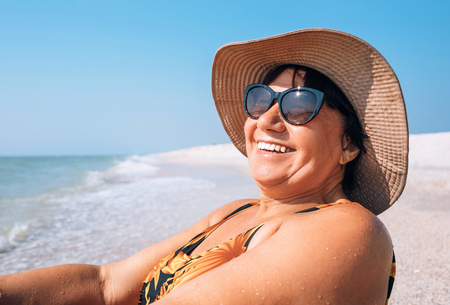 Happy smiling Elderly womanr woman in sunglasses and big hat takes sun bath on seasideの写真素材