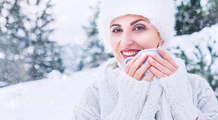 Smiling woman in cozy knitwear and fur hat with cup of hot drink in winter forest medium shot. Winter fashion concept.の写真素材