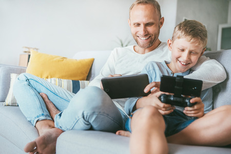 Father and son game players fun sit together at home on cozy sofa, playing the games using the tablet and game pad.の写真素材
