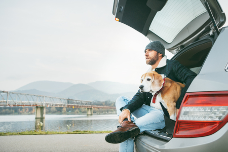Breaded man in warm casual autumn outfit siting with beagle in car trunk. Traveling with pets concept.の写真素材