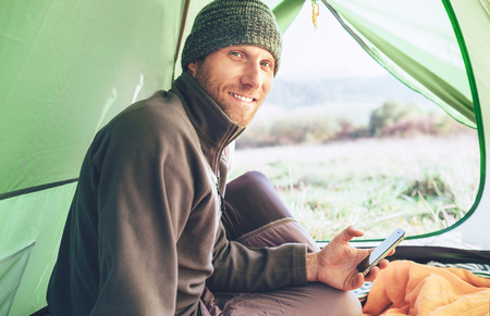 Bearded traveler warm dressed Man uses his mobile phone sitting in the camping tent and smiles looking in camera.の写真素材