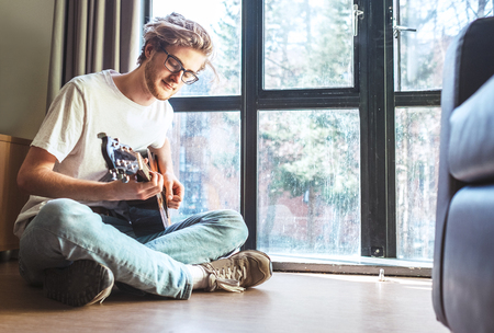 Middle shot of young man plays on guitar sitting on the floor in living room.の写真素材