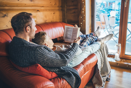 Father and son reading book together lying on the cozy sofa in warm country house. Reading to kids conceptual image.の写真素材