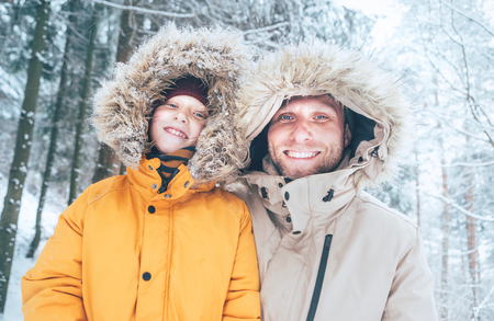 Father and son dressed in Warm Hooded Casual Parka Jacket Outerwear walking in snowy forest cheerful smiling faces portrait. Father and son relatives and winter outfit concept image.の写真素材