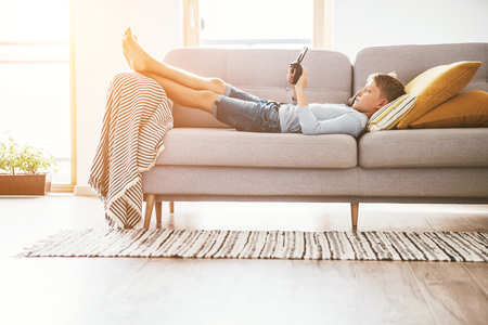 Boy plays with electronic devices - gamepad connected with smartphone lying on the cozy sofa in the home living room.の写真素材