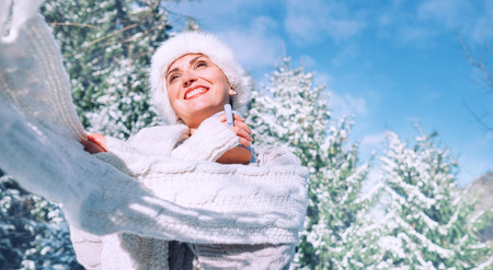 Happy smiling woman portrait in knitwear and fur hat enjoying with cup hot tea at sunny winter day during snowy forest walk.の写真素材