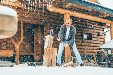 Man chopping wood on snowy yard for a house fireplace with heavy snowflakes background . Winter countryside holidays concept imageの写真素材
