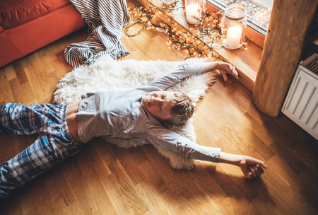 Boy stretching himself lying on floor on sheepskin and looking in window in cozy home atmosphere. Peaceful Lazy moments in cozy home concept image.の写真素材
