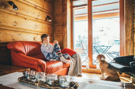 Boy sitting on cozy couch at living room and his beagle dog watching in wide window in cozy home atmosphere. Peaceful moments of cozy home concept image.の写真素材