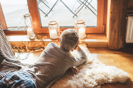 Boy lying on floor on sheepskin and looking in window in cozy home atmosphere. Peaceful moments of cozy home concept image.の写真素材