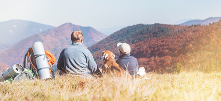 Father and son travelers with their beagle dog sit together in mountain valley with beautiful hills view. Trekking with kids and pets concept image.の写真素材