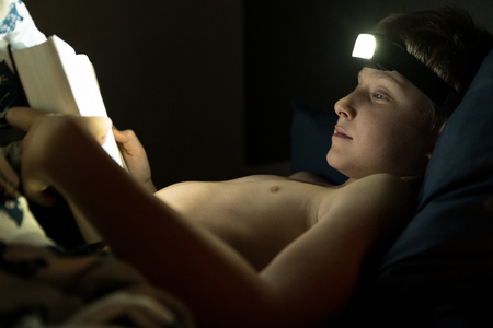 Reading in bed at the night concept photo. Little reader boy uses the Head Torch for a book reading in bedの写真素材