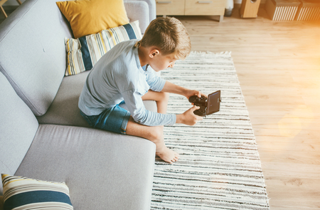 Preteen boy losted in computer game. Sits at home on coze sofa, uses gamepad with smartphoneの写真素材
