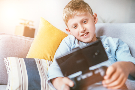 Preteen boy plays with smartphone connected with gamepad lying on the cozy sofa in the home living roomの写真素材