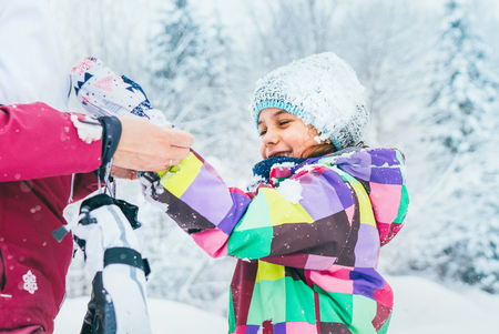 Mother helping her little daughter wearing warm gloves during snowy forest walking.の写真素材