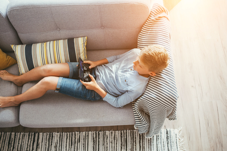 Boy laying on sofa plays with electronic devices - gamepad connected with smartphoneの写真素材