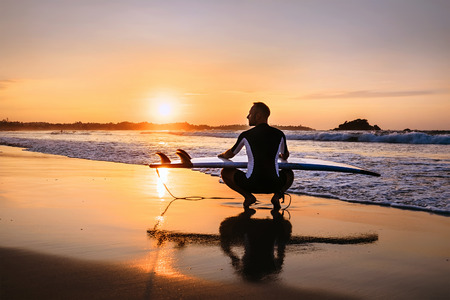 Surfer with surfboard sits on ocean surf line at sunset timeの写真素材
