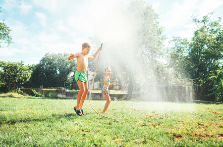Hot summer days activity: happy little girl jumps under water, when brother pours her from garden hoseの写真素材
