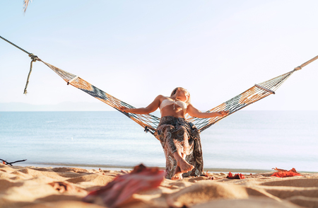 Woman swinging in hammock on sandy ocean beach gracefully bending her body enjoying sunrise lightの写真素材