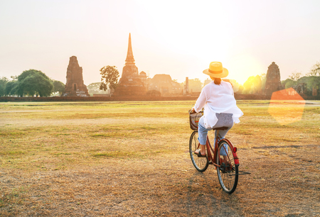 Young female weared light summer clothes and hat riding bicycle near Ayutthaya historical park in Thailand covered with morning sunlightの写真素材