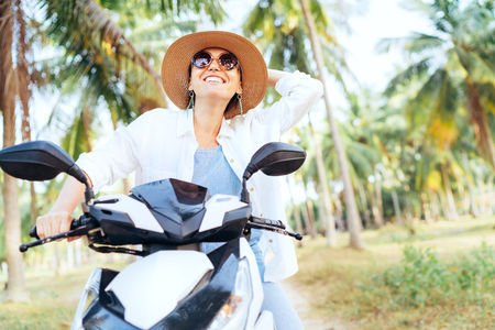 Happy smiling traveller woman riding Scooter motorbike under palm trees. Warm countries vacation concept image.の写真素材