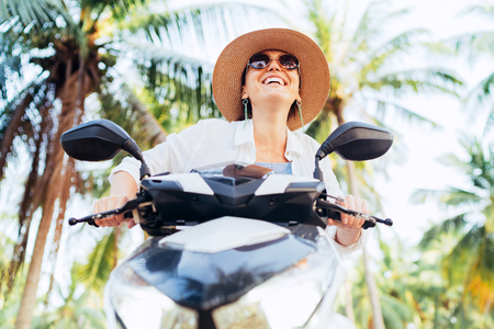 Happy smiling traveller woman riding Scooter motorbike under palm trees. Warm countries vacation concept image.の写真素材