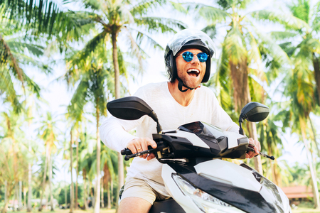 Happy smiling and screaming male tourist in helmet and sunglasses riding motorbike scooter during his tropical vacation under palm trees