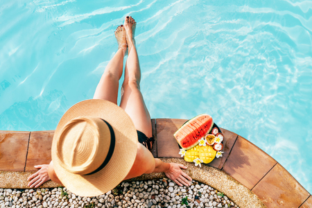 Woman in straw hat sitting on swimming pool side  with plate of tropical fruits- camera top viewの写真素材