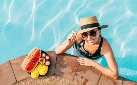Smiling woman in straw hat in sunglasses swimming in pool and enjoying fresh tropical fruitsの写真素材