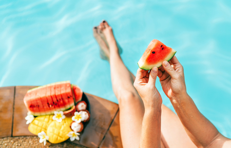 Woman take in hands little watermelon piece, sitting on the pool edge and enjoing fresh fruitsの写真素材