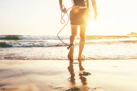 Young man Surfer taking surfboard and comming with long surf board to waves on the evening sunset sky background. Close up legs active holidays spending concept.  の写真素材