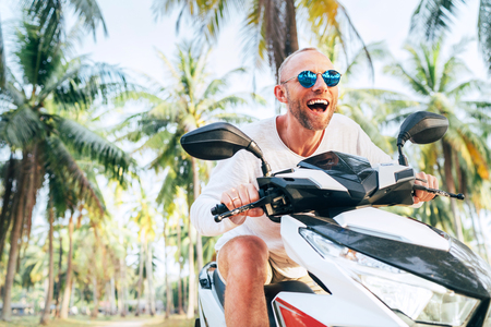 Happy smiling male tourist in sunglasses riding motorbike scooter during his tropical vacation under palm treesの写真素材