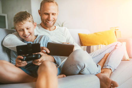 Father and son game players funs sit together at home on cozy sofa, using the tablet and gamepadの写真素材